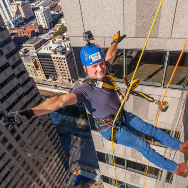 The image shows a man rappelling down the side of a tall building. He is wearing a helmet, safety harness, and gloves. The man is smiling and appears to be enjoying the experience. The background shows other buildings and the city below.